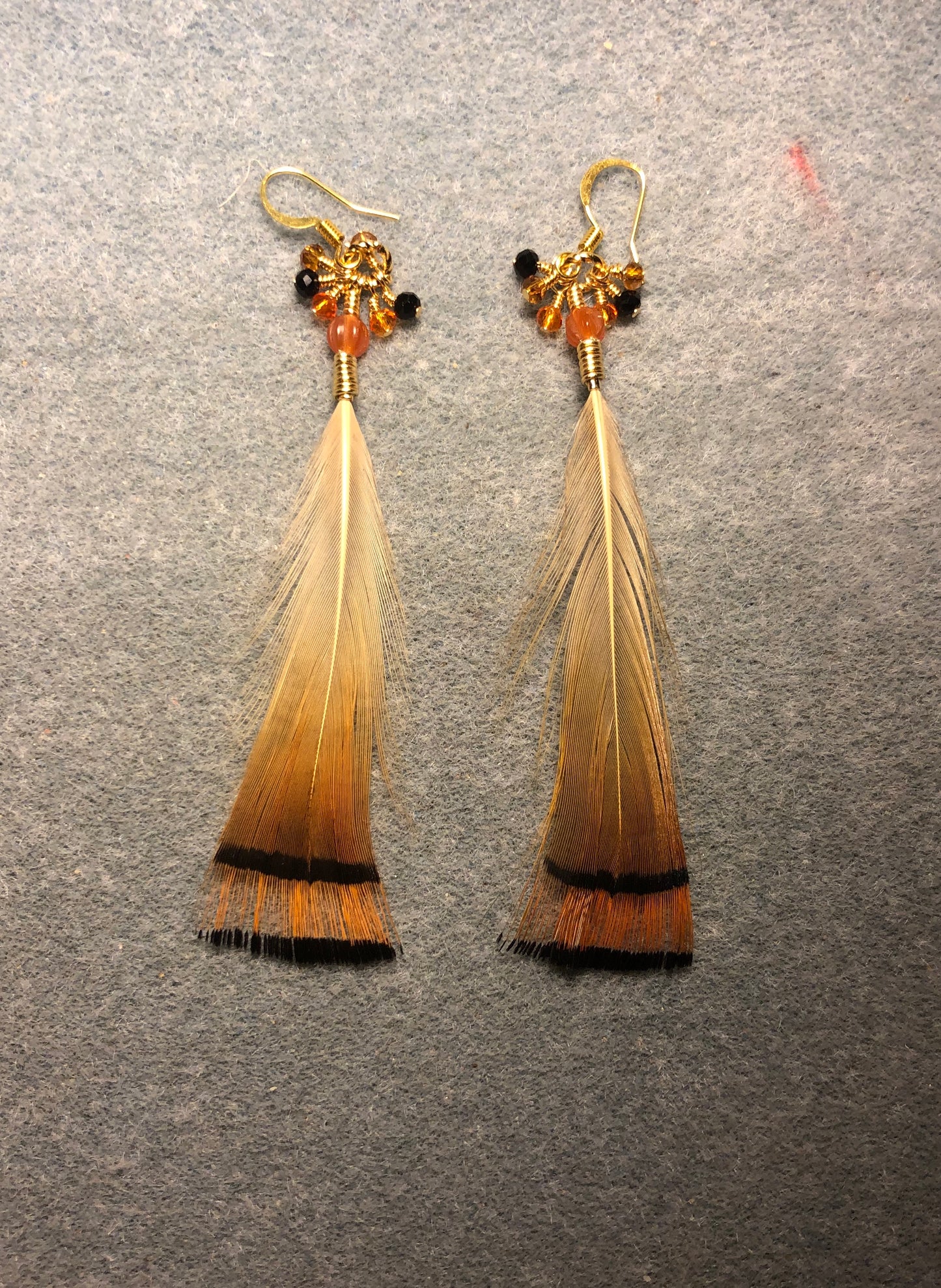 Golden pheasant tippet feather earrings adorned with orange Czech glass melon beads and tiny dangling orange, brown, and black Chinese crystal beads.
